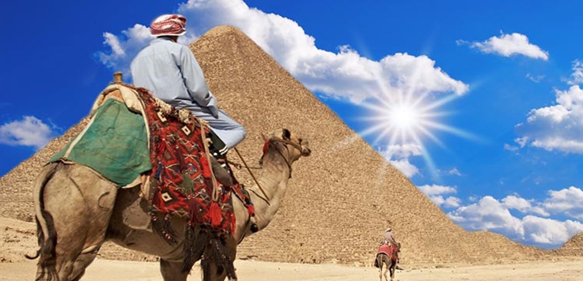 Una persona che cavalca un cammello con la Grande Piramide di Giza sullo sfondo sotto un cielo soleggiato.