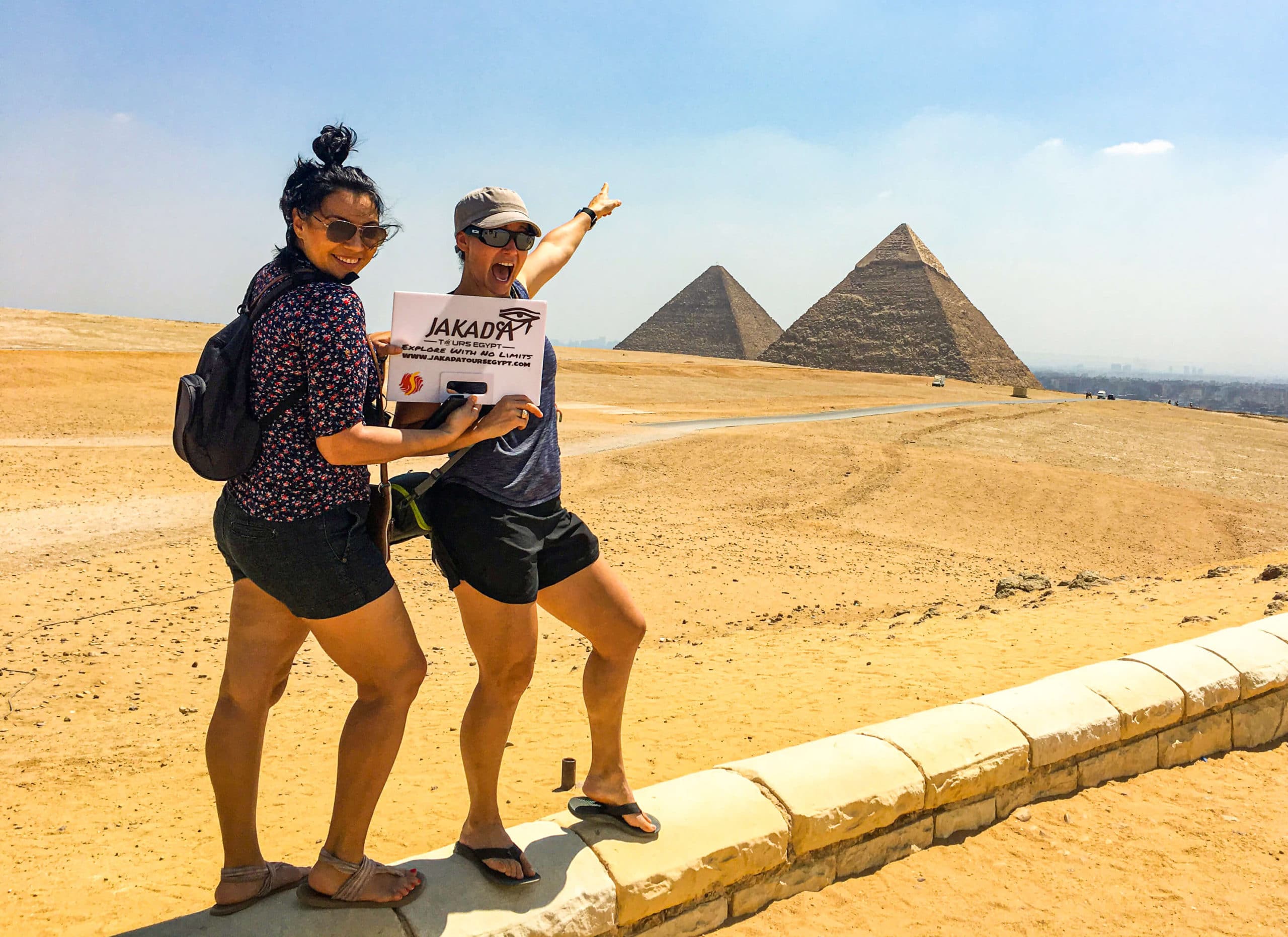 Two women posing with the Giza pyramids in the background, holding a 'Jakaby Tours Egypt' sign.