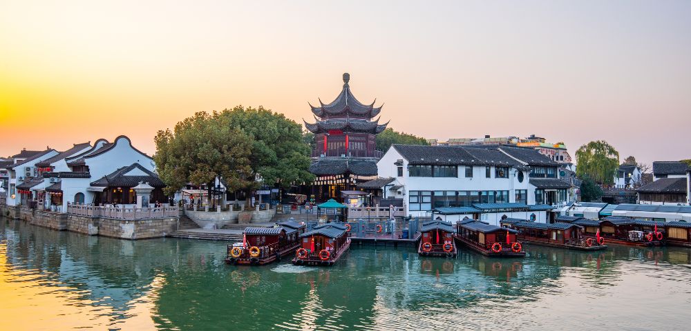 A vibrant city view in China with traditional architecture and boats on a canal at sunset.
